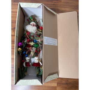 May include: A decorative Santa Claus figurine with a white beard, wearing a green and red outfit adorned with pine branches, berries, and small colorful ornaments. The figurine is placed inside an open cardboard box with a product information label.