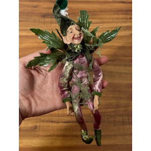 Peut inclure: Figurine de fée fantaisiste avec un visage souriant, portant un chapeau vert avec un accent blanc et un costume rose et or. La fée a des accents de feuilles vertes sur les épaules et autour de la taille, et des chaussures vertes. La figurine mesure environ 15 cm de haut.