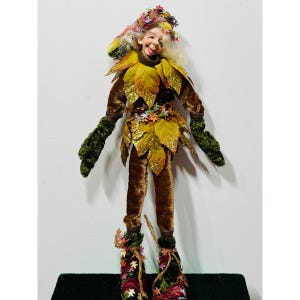 Puede incluir: Una muñeca caprichosa con temática otoñal con una cara sonriente, vestida con un atuendo adornado con hojas. La muñeca presenta un traje de terciopelo marrón, guantes verdes y detalles en forma de hoja. El sombrero de la muñeca está decorado con hojas y flores coloridas.