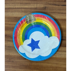 May include: A round paper plate with a rainbow design, a white cloud, and a blue star. The plate has a blue border and is wrapped in clear plastic. The plate is on a wooden surface.