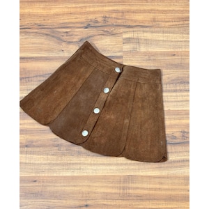 May include: A brown suede skirt with a button-up front. The skirt has a unique petal-like design and is made of a soft, textured material. The skirt is a warm brown color and is a vintage style.