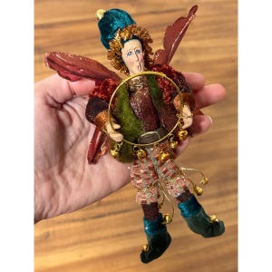 Puede incluir: Una caprichosa figura de hada con un sombrero de terciopelo verde azulado, adornos dorados y alas de color marrón rojizo. El hada viste un atuendo verde y marrón con pantalones estampados y sostiene un aro dorado con campanillas colgantes.