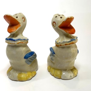 May include: A pair of vintage ceramic duck salt and pepper shakers. The ducks have open orange beaks, white bodies, and are adorned with blue and yellow accents. Each shaker has multiple holes on top.