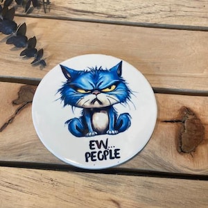 May include: A white ceramic coaster with a blue cat cartoon illustration. The cat has a grumpy expression and is sitting down. The text "EW... PEOPLE" is printed below the cat.