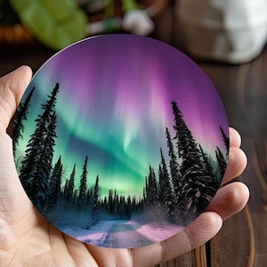 May include: A round coaster featuring a photograph of a snowy forest with a road leading through it. The sky is a vibrant purple and green, with the aurora borealis shining brightly overhead.