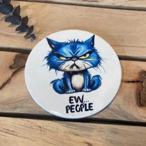 May include: A round white ceramic coaster featuring a cartoon illustration of a grumpy blue cat with yellow eyes. Below the cat, the text reads "EW... PEOPLE" in bold black lettering. The design is centered on the smooth, circular surface.