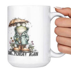 May include: White ceramic mug featuring a whimsical illustration of a frog wearing a coat and holding an umbrella. The frog is in a rainy scene with the text "Dang...Monday Again". The mug has a large handle.