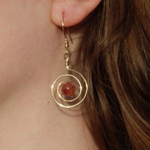 Puede incluir: Pendientes en espiral dorados con una cuenta central de color marrón rojizo. Los pendientes están envueltos en alambre y cuelgan de un gancho simple. Un accesorio único y llamativo.