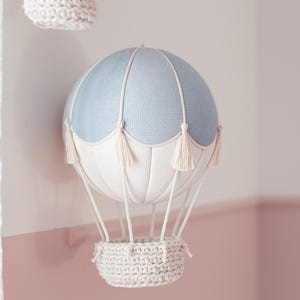 Op de afbeelding: Een lichtblauwe en witte luchtballon met een witte gehaakte mand die aan het plafond hangt. De ballon heeft witte kwastjes aan de onderkant.