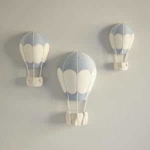 May include: Three decorative hot air balloon wall hangings. Each balloon features a light blue top and white panels, with a woven basket base. The balloons are suspended by white cords, creating a whimsical nursery or children's room decor.
