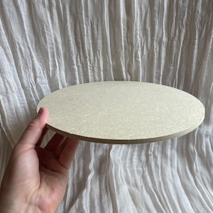 May include: A round, light beige wooden board. The board is held by a hand against a white, textured background. The board appears to be approximately 30 cm in diameter.