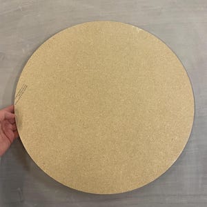 May include: A large, circular, unfinished wooden craft board. The board is a light tan colour and appears to be made of particle board or similar material. The board is held by a hand.