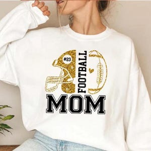 May include: White sweatshirt with a gold glitter football helmet and football graphic. The text "FOOTBALL MOM" is in black block letters. The helmet has the number "23" on it.
