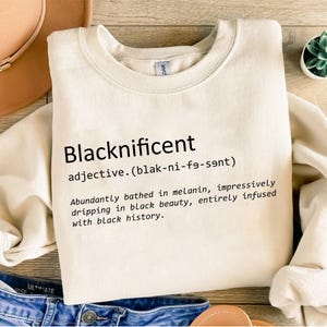 May include: Cream-colored sweatshirt with the word "Blacknificent" printed in black, along with a definition. The definition describes the term as being "bathed in melanin" and "infused with black history."