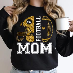 May include: Black sweatshirt with a gold glitter football helmet and football graphic. The text reads "FOOTBALL MOM" in white block letters. The helmet has the number "#23" on it. A white mug is held in the hand.