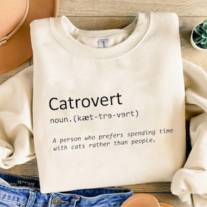 May include: Cream-colored sweatshirt with the word "Catrovert" printed in black, along with its pronunciation and definition: "A person who prefers spending time with cats rather than people."