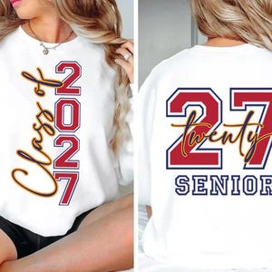 May include: White sweatshirt with "Class of 2027" and "2027" in red and gold lettering on the front. The back of the sweatshirt features "27 Twenty SENIOR" in red, white, and gold lettering. A graduation-themed apparel item.