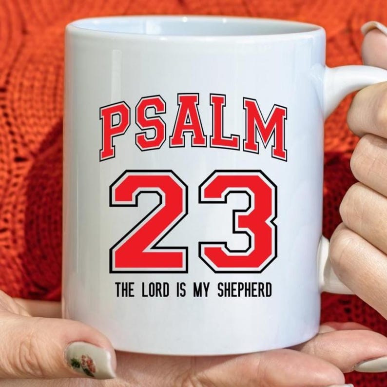 May include: White ceramic mug with the text "PSALM 23" in red block letters with a black outline. Below the number "23" is the text "THE LORD IS MY SHEPHERD" in black. The mug has a white handle.