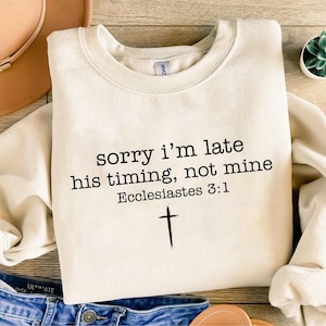 以下が含まれることがあります： クリーム色のスウェットシャツ。黒字で「sorry i'm late his timing, not mine Ecclesiastes 3:1」と書かれています。テキストの下には黒い十字架があります。スウェットシャツは明るい色の表面に折りたたまれています。