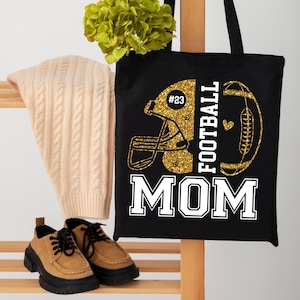 May include: Black tote bag with a gold glitter football helmet and football graphic. The text "FOOTBALL MOM" is in white block letters. A beige cable-knit sweater and brown shoes with black soles are also visible.