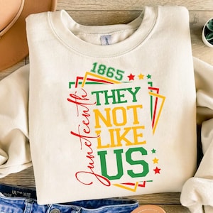 May include: Cream-colored sweatshirt with the text "Juneteenth" in red script and the phrase "1865 THEY NOT LIKE US" in green, yellow, and red. The design includes stars and geometric shapes.
