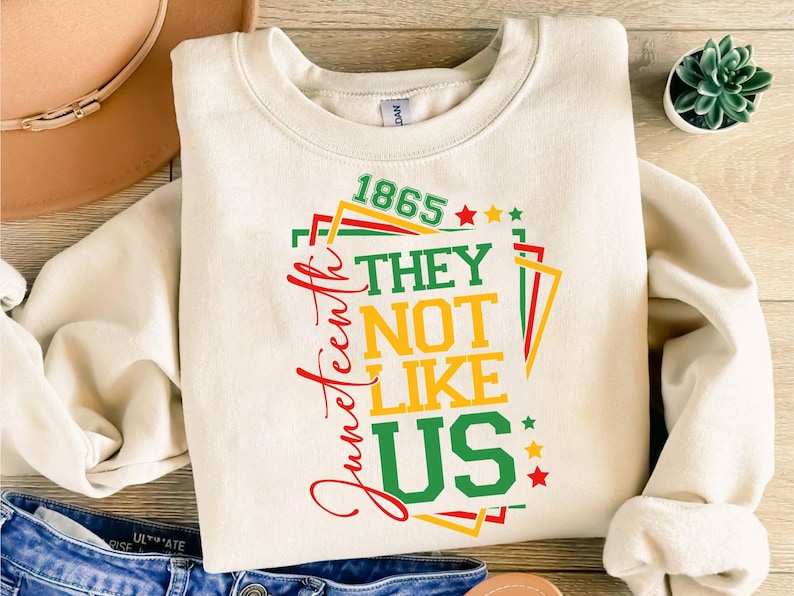 May include: Cream-colored sweatshirt with the text "Juneteenth" in red script and the phrase "1865 THEY NOT LIKE US" in green, yellow, and red. The design includes stars and geometric shapes.