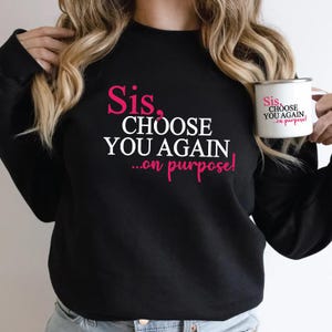 Könnte beinhalten: Schwarzes Sweatshirt mit dem Text "Sis, CHOOSE YOU AGAIN ...on purpose!" in Rosa und Weiß. Eine weiße Tasse mit dem gleichen Text wird in der Hand gehalten.