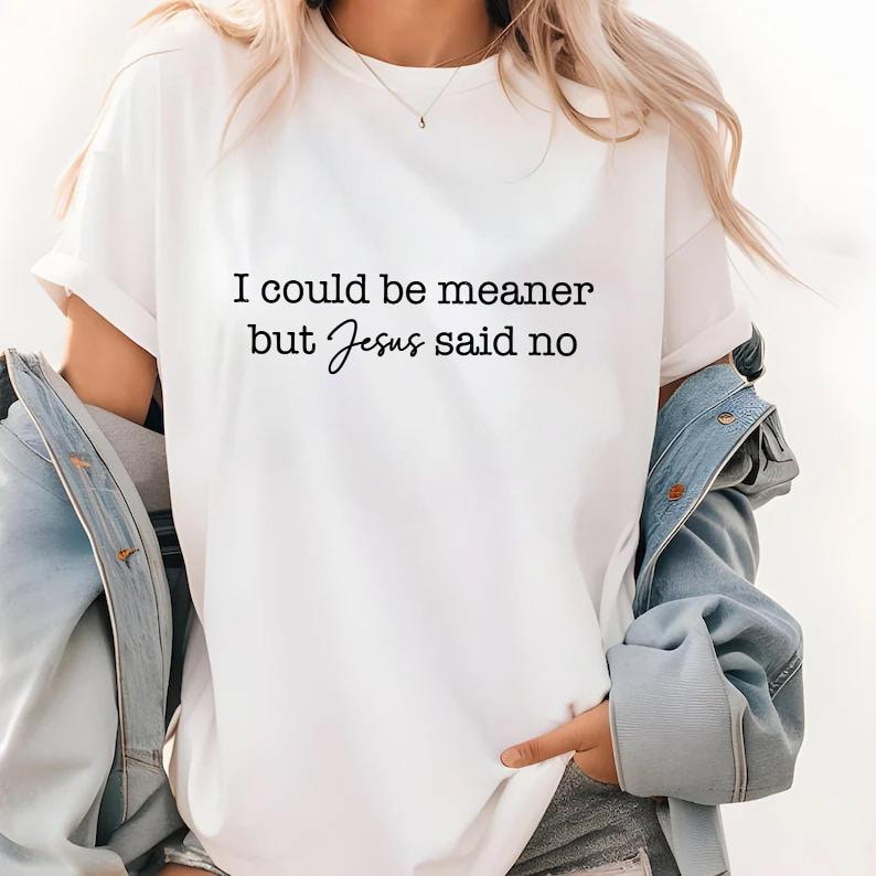 May include: White t-shirt with the text "I could be meaner but Jesus said no" in black script. The shirt is a casual style, with rolled-up sleeves. A denim jacket is draped over the shoulders.