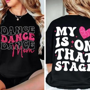 Puede incluir: Camiseta negra con las palabras "DANCE DANCE DANCE Mom" en letras blancas y purpurina rosa en la parte delantera. La parte trasera de la camiseta dice "MY IS ON THAT STAGE" con un corazón de purpurina rosa.
