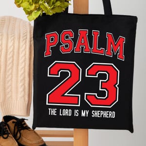 May include: Black tote bag with the words "PSALM 23" in red block letters with a white outline. Below the number "23" is the text "THE LORD IS MY SHEPHERD" in white. The bag has black handles.