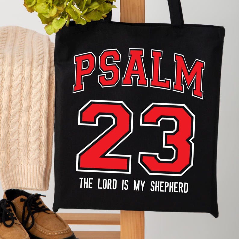 May include: Black tote bag with the words "PSALM 23" in red block letters with a white outline. Below the number "23" is the text "THE LORD IS MY SHEPHERD" in white. The bag has black handles.