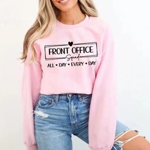 Puede incluir: Una sudadera rosa claro con el texto "FRONT OFFICE Squad" en un recuadro negro, con "ALL DAY EVERY DAY" debajo. La sudadera se combina con unos vaqueros azules.