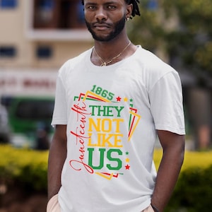 May include: White t-shirt with a colorful graphic design. The design includes the text "1865", "Juneteenth", "THEY", "NOT", "LIKE", and "US" in red, green, and yellow. The shirt is worn by a person.
