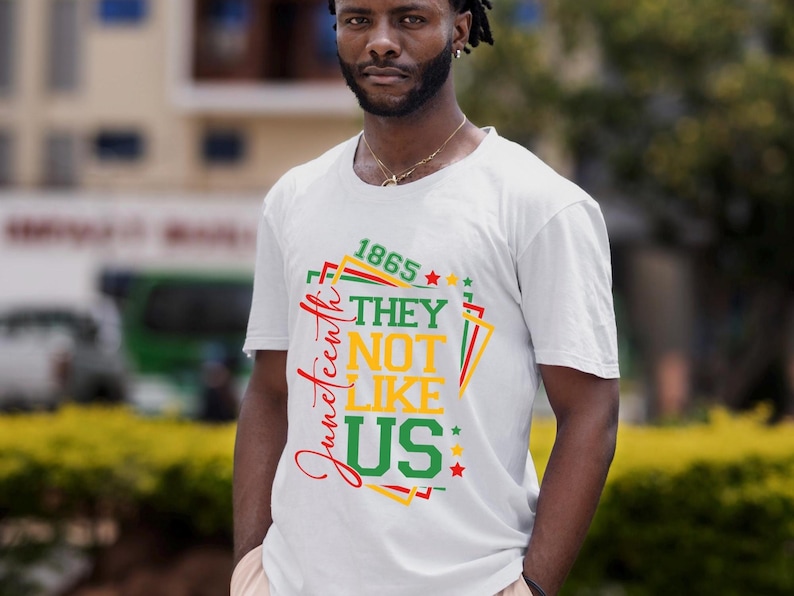 May include: White t-shirt with a colorful graphic design. The design includes the text "1865", "Juneteenth", "THEY", "NOT", "LIKE", and "US" in red, green, and yellow. The shirt is worn by a person.