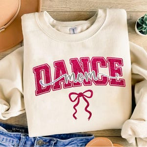 May include: Cream-colored sweatshirt with the words "DANCE mom" in a glittery pink and silver design. A pink bow graphic is below the text. The sweatshirt is a casual style.