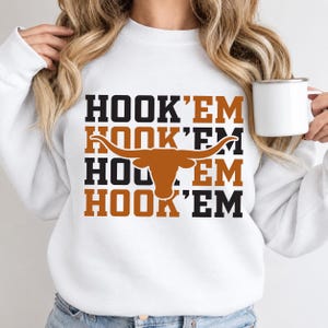 Op de afbeelding: Een wit sweatshirt met lange mouwen met de tekst HOOK 'EM vier keer herhaald in afwisselende kleuren zwart en gebrand oranje. Een groot silhouet van een longhorn-rund in gebrand oranje staat centraal over de tekst en vervangt de middelste letters.
