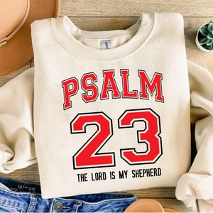 May include: Cream-colored sweatshirt with the text "PSALM 23" in red block letters with a black outline. Below the numbers "23" is the phrase "THE LORD IS MY SHEPHERD" in black. The sweatshirt is a casual, comfortable style.