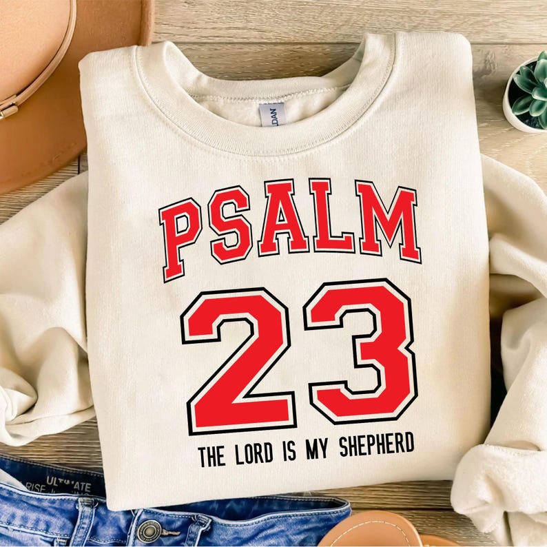 May include: Cream-colored sweatshirt with the text "PSALM 23" in red block letters with a black outline. Below the numbers "23" is the phrase "THE LORD IS MY SHEPHERD" in black. The sweatshirt is a casual, comfortable style.