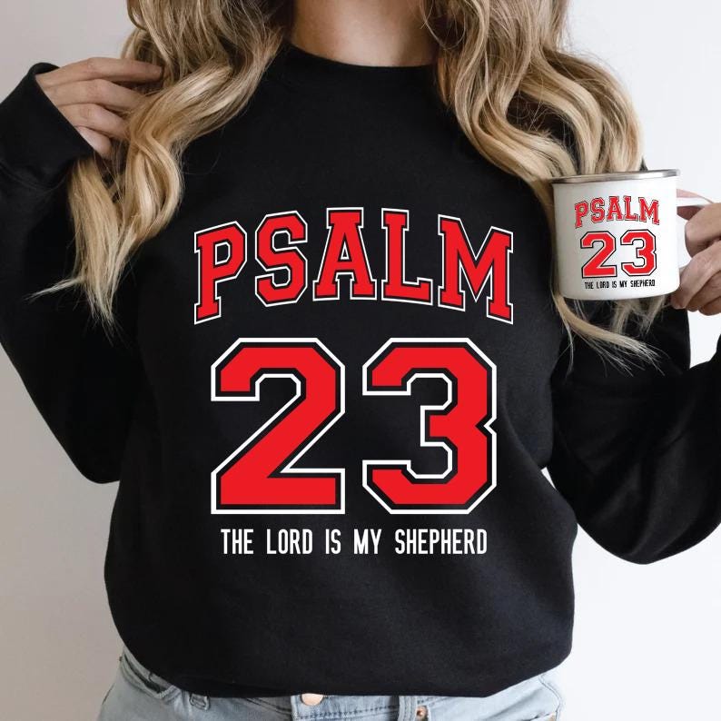 May include: Black sweatshirt with the text "PSALM 23" in red and white. A white mug with the same text is held in the hand. The phrase "THE LORD IS MY SHEPHERD" is printed below the numbers.