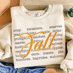 May include: Cream-colored sweatshirt with the word "Fall" in glittery orange script. Surrounding the word are black text phrases like "cozy," "sweaters," "pumpkin patches," and "bonfires," evoking autumn themes.
