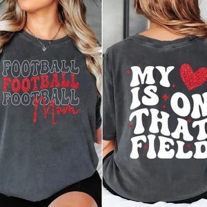 May include: Dark gray t-shirt with the words "FOOTBALL FOOTBALL FOOTBALL Mom" in white and red glitter lettering on the front. The back of the shirt says "MY HEART IS ON THAT FIELD" with a red glitter heart.