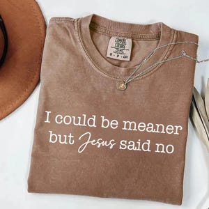 May include: A brown t-shirt with the text "I could be meaner but Jesus said no" in white script. A silver necklace with a small pendant rests on the shirt. The shirt is folded, with a hat visible in the background.