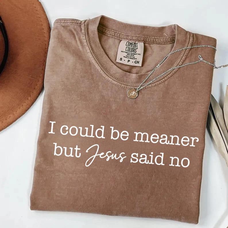 May include: A brown t-shirt with the text "I could be meaner but Jesus said no" in white script. A silver necklace with a small pendant rests on the shirt. The shirt is folded, with a hat visible in the background.