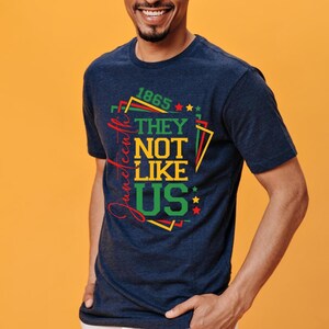May include: Navy blue t-shirt with the text "1865 They Not Like Us" in red, green, and yellow. The design includes stars and geometric shapes. The word "Juneteenth" is written vertically in red.