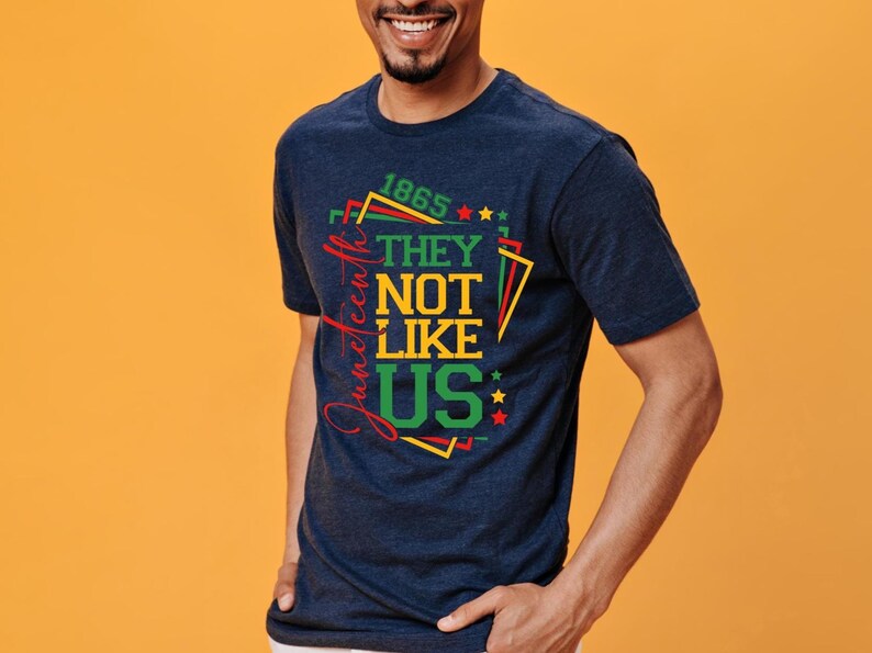 May include: Navy blue t-shirt with the text "1865 They Not Like Us" in red, green, and yellow. The design includes stars and geometric shapes. The word "Juneteenth" is written vertically in red.
