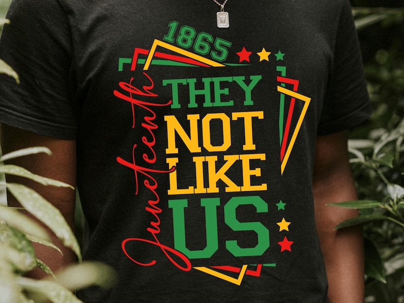 May include: Dark gray t-shirt with a colorful graphic design. The design includes the words "Juneteenth" in red script, and "1865 THEY NOT LIKE US" in yellow and green block letters, with stars and geometric shapes.