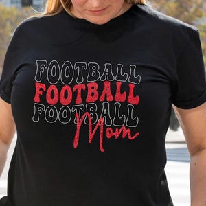 May include: Black t-shirt with the words "FOOTBALL" stacked three times, with the middle one in red glitter and outlined in white. The word "Mom" is written in red glitter below the stacked words.