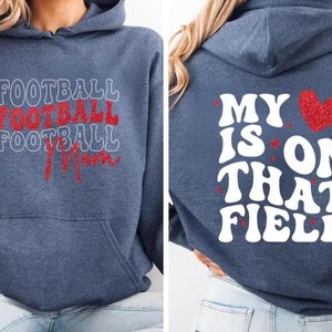May include: A navy blue hoodie with the words "FOOTBALL FOOTBALL FOOTBALL Mom" in white and red lettering on the front. The back of the hoodie has the phrase "MY HEART IS ON THAT FIELD" with a red heart graphic.