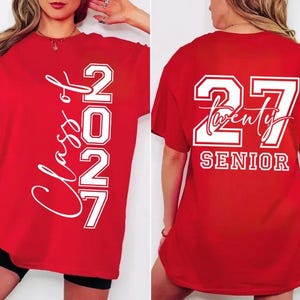 May include: Red t-shirt with white text. The front of the shirt says "Class of 2027" vertically. The back of the shirt says "27 Twenty SENIOR". The shirt is a graduation-themed apparel item.