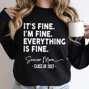 May include: A black long-sleeve sweatshirt featuring white distressed text that reads: IT'S FINE. I'M FINE. EVERYTHING IS FINE. Below this, the text reads: Senior Mom - CLASS OF 2027 -. The sweatshirt has a crew neckline and long sleeves.